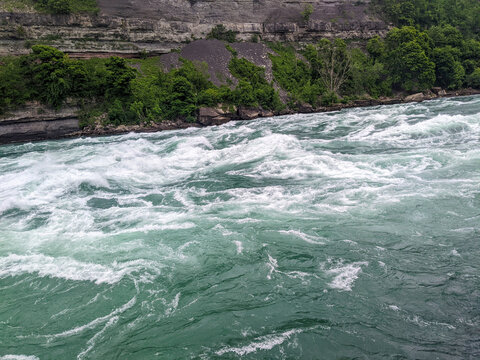 Turquoise rapids flow on the Niagara River with layered limestone from the Niagara Escarpment cliff in the background during summer.