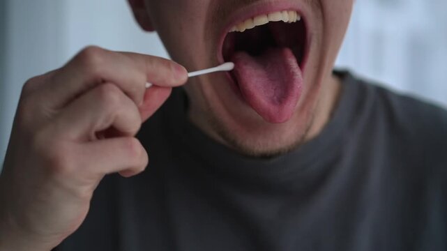A young man takes a saliva sample with a cotton swab, placing it inside his mouth to gather material for testing. The scene reflects a simple at-home procedure for DNA testing or medical diagnostics.