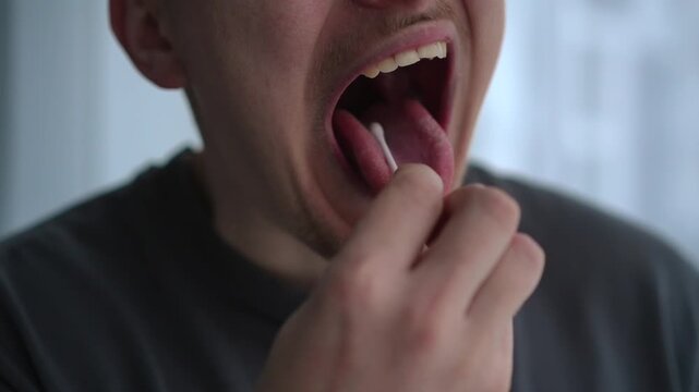 A young man collects a saliva sample using a cotton swab, opening his mouth wide in a bright room. He carefully swabs inside his cheek, suggesting a home DNA test or health analysis procedure.
