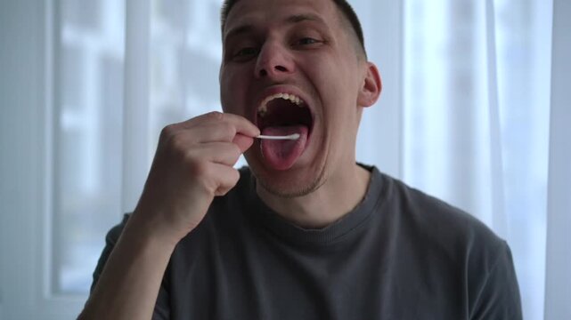 A young man collects a saliva sample using a cotton swab, opening his mouth wide in a bright room. He carefully swabs inside his cheek, suggesting a home DNA test or health analysis procedure.