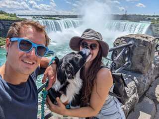 Obraz premium Happy couple pose with their dog for a selfie at Niagara Falls. The border collie kisses the woman while enjoying sunny weather and misty waterfall views on a scenic riverside overlook.