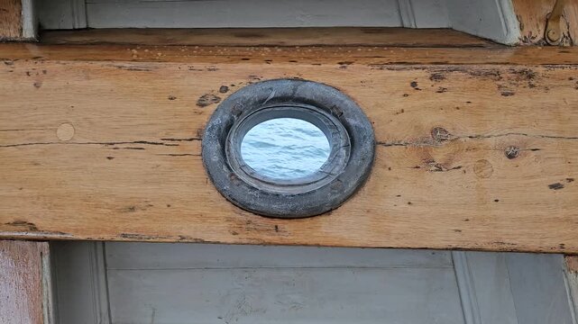 A rustic wooden boat porthole framing a calm ocean view through a round metal window on an aged ship deck, highlighting weathered wood grain, nautical character and vintage maritime texture.