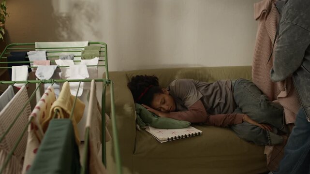 Full shot of loving Black mother covering with blanket tired teenage daughter resting on couch after doing homework, managing childcare and housework in modest apartment