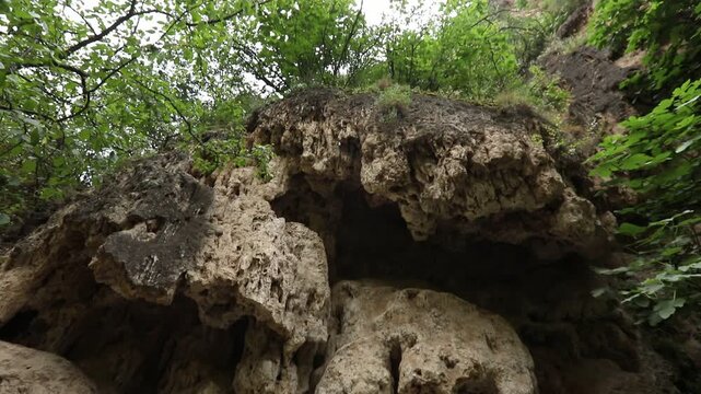 The video begins with a close-up of a highly textured porous rock overhang. The camera then slowly tilts upwards revealing a steep, rugged cliff face with green vegetation.

