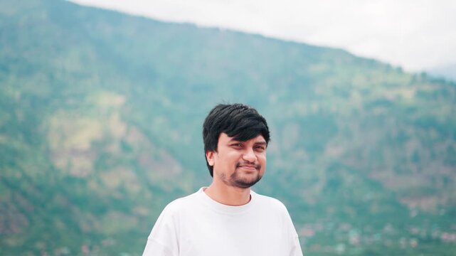 4K Portrait shot of a smiling young Indian man looking at the camera while standing in front of the Himalayan mountain peaks during the summer season at Manali in Himachal Pradesh, India. 