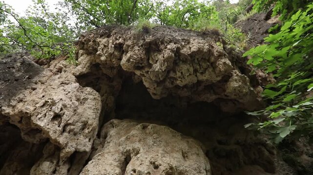 The video begins with a close-up of a highly textured porous rock overhang. The camera then slowly tilts upwards revealing a steep, rugged cliff face with green vegetation.


