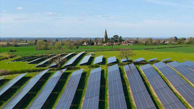 Aerial view of a large solar farm with rows of blue panels set against green fields and the village of Nailstone with its church spire under a clear sky in Nailstone, England, United Kingdom.