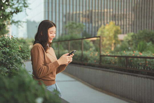 Happy young Asian woman in brown sweater using smartphone in modern urban park. Cheerful Gen Z student or digital nomad chatting on mobile phone with a smile. Concept of digital connectivity, social m