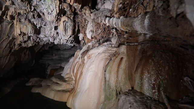 A panning shot reveals gentle water flowing over smooth, rounded rock formations inside a dark cave, some with green moss.
