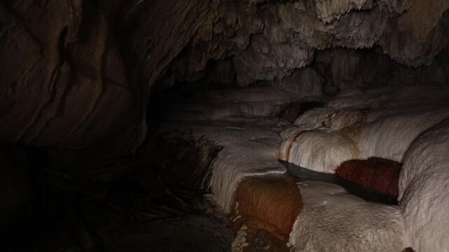 A panning shot reveals gentle water flowing over smooth, rounded rock formations inside a dark cave, some with green moss.
