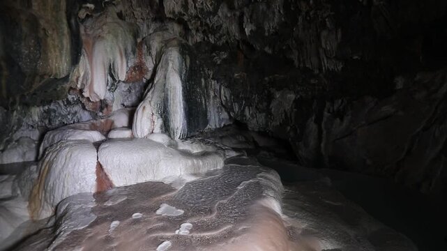 A panning shot reveals gentle water flowing over smooth, rounded rock formations inside a dark cave, some with green moss.
