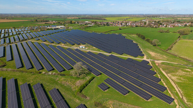 Aerial view of rows of solar panels on green hills with a village in the background under a blue sky in Nailstone, England, United Kingdom.