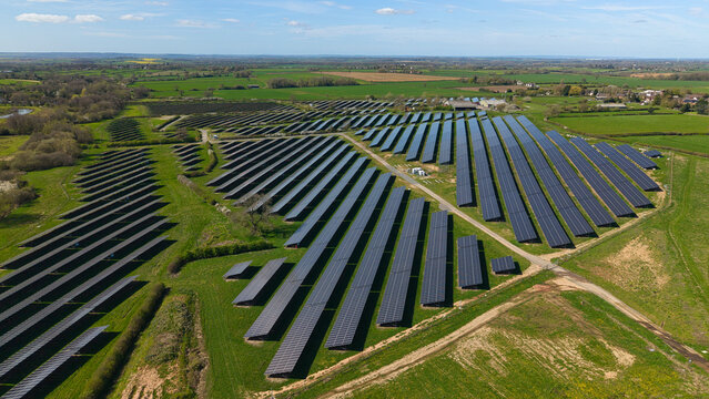 Aerial view of a large solar farm with rows of photovoltaic panels surrounded by green fields and trees under a clear blue sky in Nailstone, England, United Kingdom.