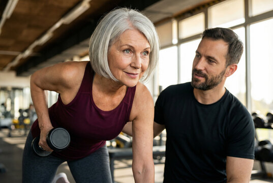 Personal trainer assisting senior woman with dumbbell row in gym