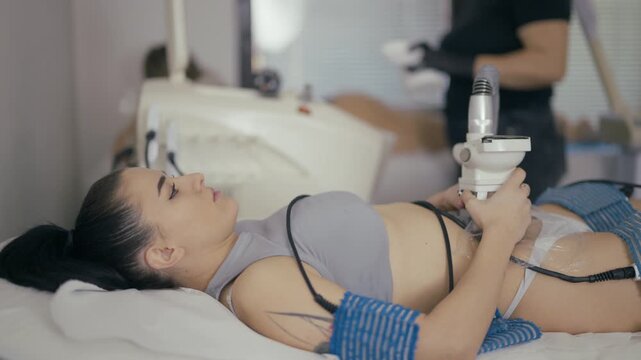 A young woman receives a non-invasive body contouring procedure at a beauty salon. The client lies calmly while a specialist administers the cosmetic treatment.