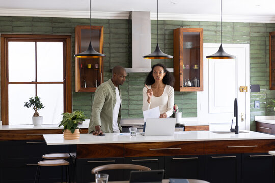 Couple reviewing papers while leaning on island with laptop, glass, plants in modern kitchen
