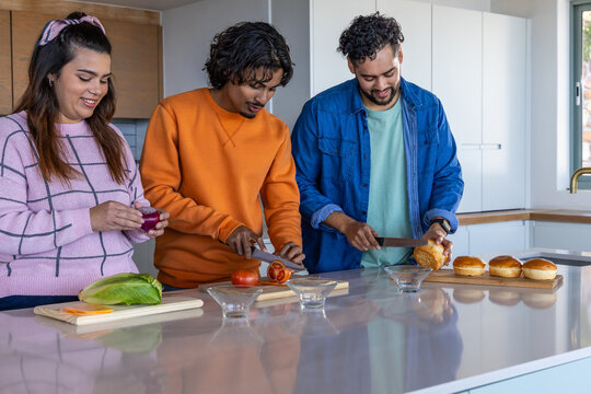Diverse friends prepping at glossy kitchen island, slicing tomato on wooden cutting boards
