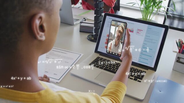 Man in yellow raising phone over laptop, math overlays appearing while reviewing docs for education