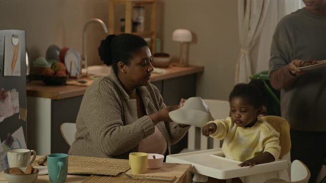 Medium long shot of Black teenage girl serving lunch to her mother tying baby bib on toddler sister sitting in high chair, gathered around small table to share modest meal together at home