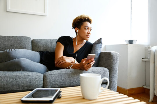 Young woman sitting on sofa with smartphone and earphones looking away at home
