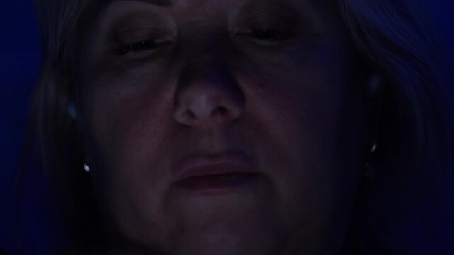 Close up face of a mature woman illuminated by a screen light in the dark. Middle aged lady scrolling news or reading messages on a gadget at nigh