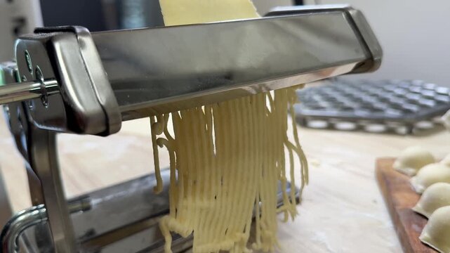 Fresh pasta strands extrude from a stainless steel pasta machine on a kitchen counter, close up food preparation scene, homemade Italian cooking mood