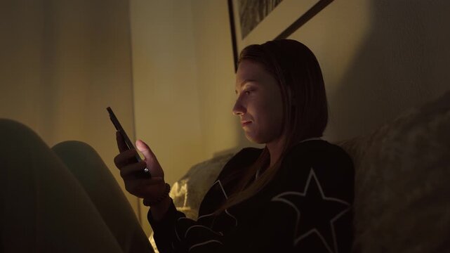 Girl doom scrolling her smartphone on a coach at night. Social network and phone addiction concept. Cozy home ambient light
