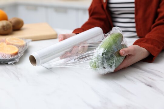 Woman wrapping fresh cucumbers in cling film at light marble table, closeup