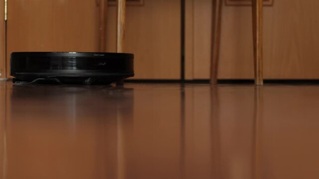 Smart robot vacuum cleaner maneuvers between the legs of wooden chairs on a glossy floor. Robotic cleaner performs a cleaning program in the dining area
