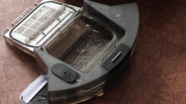 Close-up of an open robot vacuum container clogged with dust and long hair. A person uses a special brush to clean the filter and dustbin from debris
