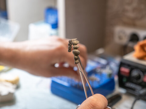 Dental bridge framework, metal crown restoration in laboratory, technician holding prosthetic teeth with tweezers, prosthodontic dentistry, precision medical technology.