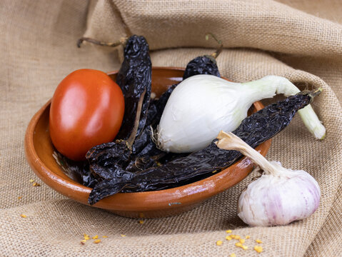 A Mexican plate with raw onions, pasilla peppers, tomatoes, and garlic, set against a textured brown background.
