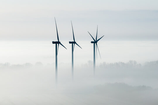 Aerial view of three wind turbines rising above a thick layer of morning fog and mist over rural fields in Wislinka, Pomeranian Voivodeship, Poland.