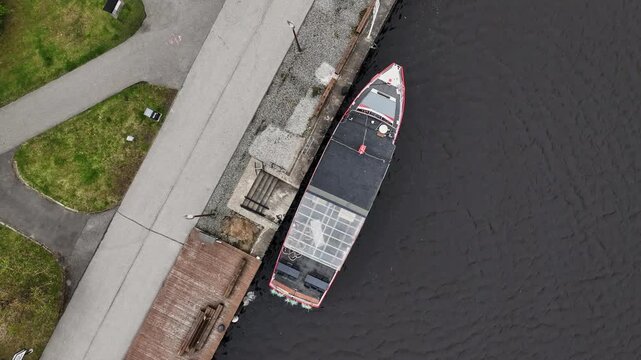 Aerial video of docked red-and-black boat with glass roof by wooden pier and stone quay. Grey pedestrian paths and green grass meet the dark Parnu River waters. P&auml;rnu, Estonia. Drone slowly moving up