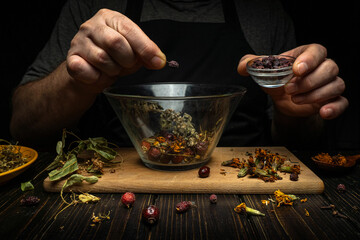 A person adds dried herbs and flowers to a glass bowl on a wooden board. They hold a small dish in their other hand. The kitchen is dimly lit, creating a focused scene © Віталій Б.