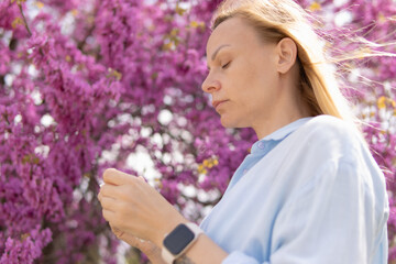 Blonde woman relaxing in a park, enjoying spring blooming pink flowers, feeling peace, wellness and serenity with eyes closed, finding comfort in nature