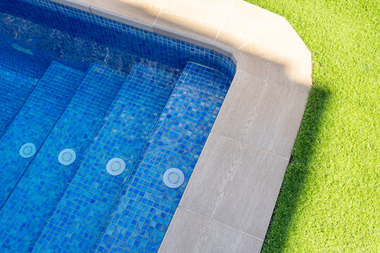 Close-up of luxury swimming pool steps with blue mosaic tiles, water jets, and clean design, surrounded by green grass - relaxation and luxury lifestyle concept