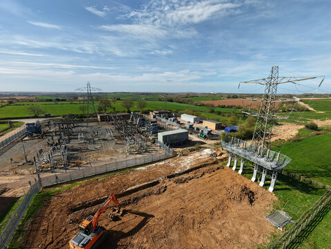 Aerial view of Coalville Substation with an orange excavator on a dirt site, electrical pylons, and transformers under a blue sky Coalville, England, United Kingdom.