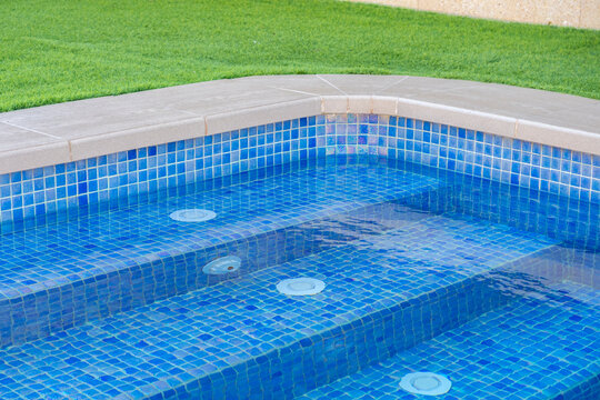 Close-up of luxury swimming pool steps with blue mosaic tiles, water jets, and clean design, surrounded by green grass - relaxation and luxury lifestyle concept