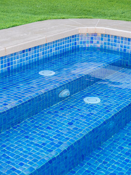 Close-up of luxury swimming pool steps with blue mosaic tiles, water jets, and clean design, surrounded by green grass - relaxation and luxury lifestyle concept