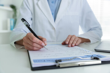Fototapeta premium Doctor in medical coat writing on clipboard at white table in clinic, closeup