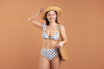 Beautiful woman wearing stylish swimsuit, hat and bag on brown background