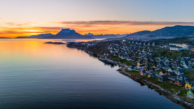 Aerial view of colorful houses along the rocky coastline of Nuuk with calm sea water and distant mountains under a vibrant sunset sky in Nuuk, Sermersooq Municipality, Greenland.