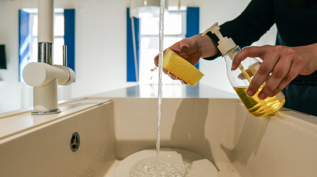 Hands holding sponge and dish soap under running water in a modern kitchen sink