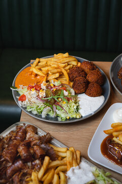A mixed restaurant plate with crispy fries, falafel, sausages, fresh salad, and creamy sauces served on a wooden table.