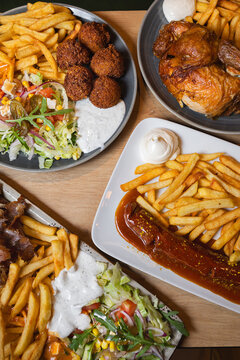 Overhead view of multiple plates with fries, sausage, roasted chicken, falafel, fresh salad, and sauces served on a wooden table.