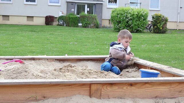 Little boy playing alone in a sandbox outdoors, focused on building and exploring with sand toys. Natural candid moment of independent play, imagination.
