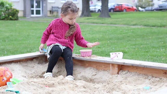 Little girl playing alone in a sandbox outdoors, focused on building shapes with sand toys. Cute candid moment of independent play, creativity, and early childhood development in a calm residential en