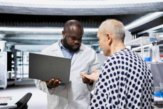 Black scientist observes patient reactions during a drug trial in a healthcare institute, recording data and monitoring vitals to ensure safe dosage levels for progress in medicine.