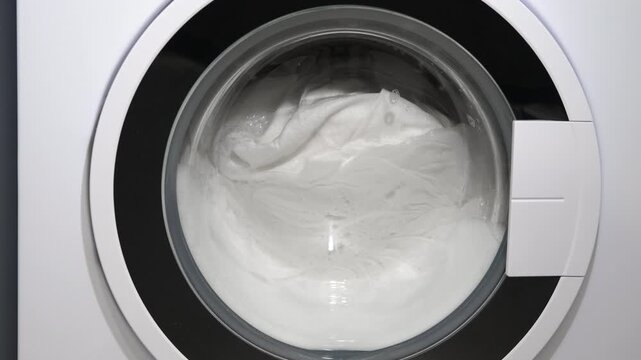 Washing machine drum spinning with white laundry. Close-up of a washing machine door showing the stainless steel drum spinning and tumbling clean white clothes in soapy water during a wash cycle